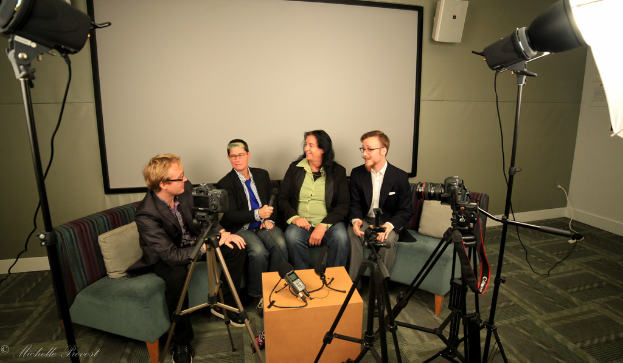 Eine Gruppe von drei Personen sitzt auf einem Sofa und gibt ein Interview, wobei eine Person links in einem schwarzen Mantel und Hose, eine Person in der Mitte in einem Mantel und blauer Krawatte mit einem Mikrofon und eine strahlende Person rechts zu sehen ist. Hinter ihnen befindet sich ein Projektionsschirm, ein Mikrofonrecorder auf dem Tisch in der Mitte und Kameras mit großen Scheinwerfern auf der rechten und linken Seite.