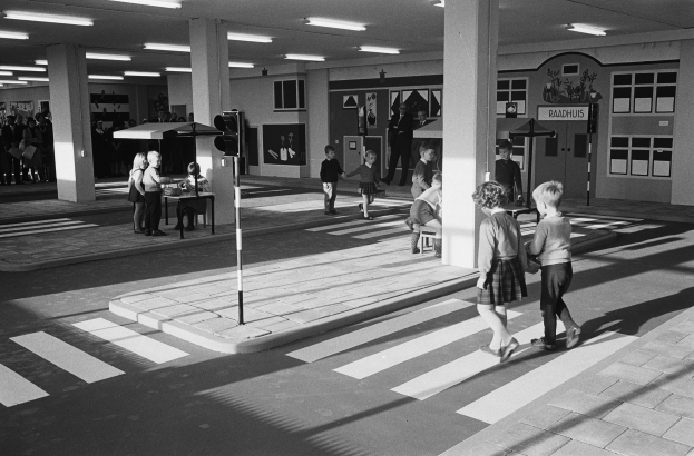 Schwarzes und weißes Foto von Kindern, die eine Straße an einer Ampel überqueren, mit Fußgängern in der Nähe, einem Verkehrssignalmast im Vordergrund und strukturellen Säulen mit Deckenleuchten im Hintergrund.