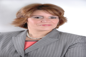 Eine professionelle Frau in einem marineblauen Anzug mit weißem Hemd und gemustertem Schlips posiert selbstbewusst vor einem weißen Hintergrund, ihre Haltung aufrecht und ihr Blick direkt.