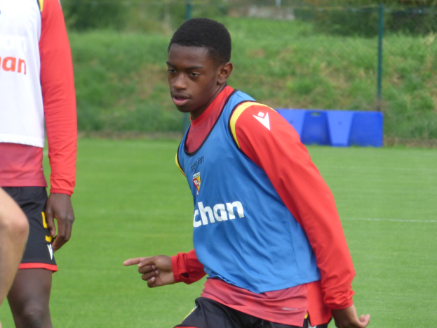 Ein junger Mann in einem blauen Hemd und schwarzen Shorts spielt Fußball auf einem grünen Feld, das von einem Zaun, Pflanzen und Bäumen umgeben ist, während eines Trainings.