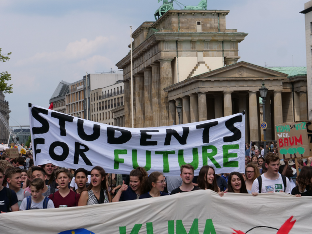 Gruppe von Schülern marschiert in Berlin mit einer bunt bemalten "Students for Future"-Schlagzeile vor einem Hintergrund aus Gebäuden, Bäumen und Himmel.
