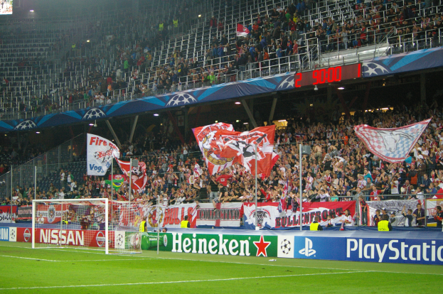 Ein Fußballspiel wird in einem gut beleuchteten Stadion mit einer großen Zuschauermenge, grünem Rasen, zentralem Torpfosten mit Bannern und einem Bildschirm in der oberen linken Ecke gespielt.