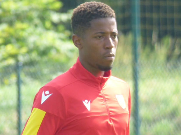 Ein junger Mann in einem roten und gelben Fußballtrikot steht auf einem Feld mit einem Zaun und Bäumen im Hintergrund und sieht konzentriert aus.