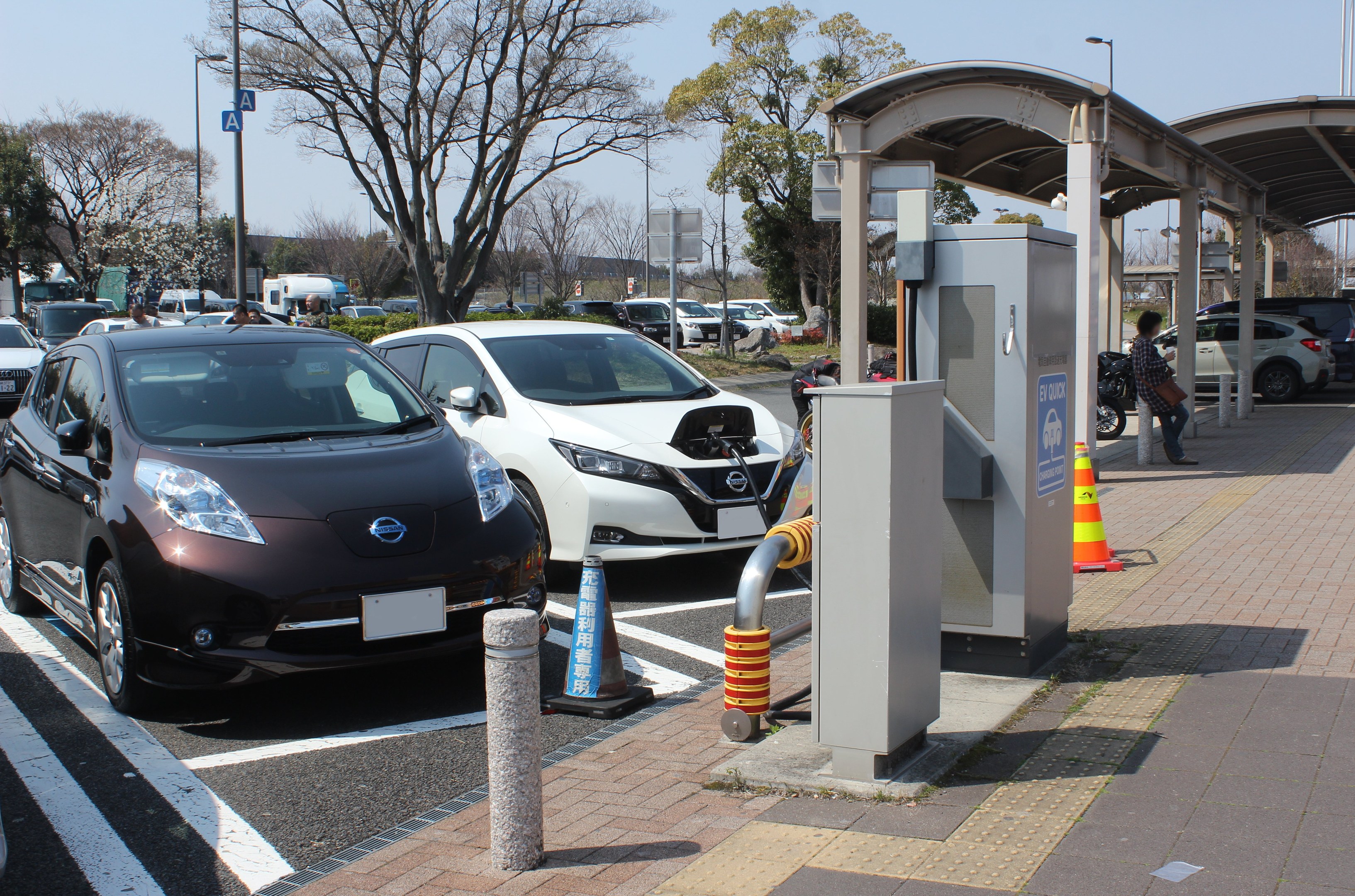 Elektroauto-Ladestation in Japan mit fahrenden Autos auf der Straße, einer Person auf dem Gehweg und umgeben von Infrastruktur gegen einen Himmelhintergrund.
