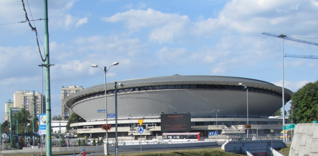 Gro├če Stadion mit starkem Verkehr davor, umgeben von Geb├Ąuden, Stra├čeninfrastruktur, Fahrzeugen, Vegetation und einem bew├Âlktem Himmel.