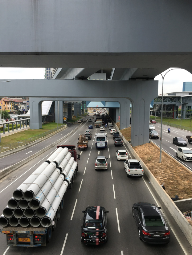 Eine stark befahrene Autobahn mit Schwerlastverkehr unter einer Brücke, umgeben von Gras, Bäumen, Gebäuden, Polen und Straßenlaternen unter einem klaren Himmel.
