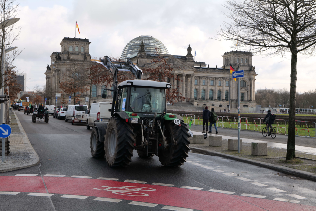 Ein Traktor fährt eine Straße vor dem Reichstaggebäude in Berlin, Deutschland, entlang, gesäumt von Bäumen, Laternenmasten und Schildern, und Menschen gehen und fahren Fahrräder auf dem Gehweg, unter einem bewölkten Himmel mit Fahnen, die das Gebäude schmücken.