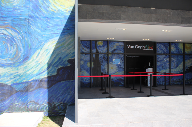 Der Eingang eines Gebäudes zeigt ein großes Wandgemälde, ein Schild mit der Aufschrift "Van Gogh Alive", Absperrbögen mit Bändern, Gras auf dem Boden und Lampen an der Decke.