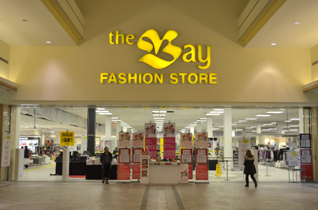 Verkaufsfläche eines Bay Fashion Retail-Shops in einem Einkaufszentrum, mit vorbeigehenden Einkäufern, Texttafeln an den Wänden, tragenden Säulen, Deckenbeleuchtung und Hintergrundauslagen.