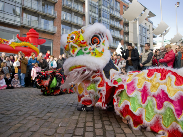 Ein lebendiges chinesisches Neujahrsfest in Amsterdam mit einer Löwen-Tanzvorstellung vor einem Publikum, das Kameras hält, vor einer Kulisse aus Gebäuden, Laternenmasten und einem klaren blauen Himmel.