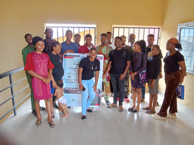 Eine Gruppe von Menschen, die drinnen zusammenstehen neben einer Tafel, mit einem Geländer auf der linken Seite, Fenstern und einer Wand im Hintergrund.