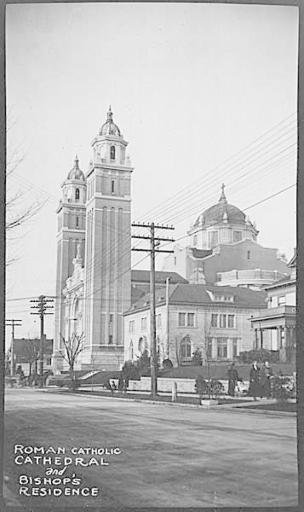 Ein Schwarz-Weiß-Foto einer römisch-katholischen Kathedrale und eines Bischofssitzes mit Gebäuden, Bäumen, Pfählen, Drähten, Menschen und einer Straße im Vordergrund und dem Himmel im Hintergrund, mit Text am unteren Bildrand.