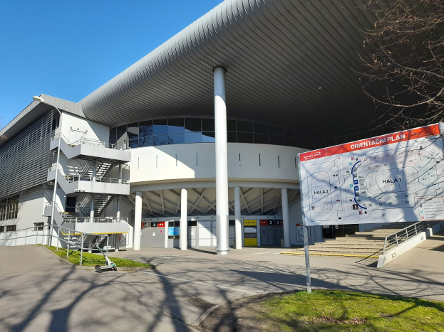 Ein großes Gebäude mit vielen Fenstern, Säulen und Treppen, das als Eingang zum Olympiagelände in London dient, mit einem Wagen, Gras, einer Straße, Bäumen und Himmel im Hintergrund.