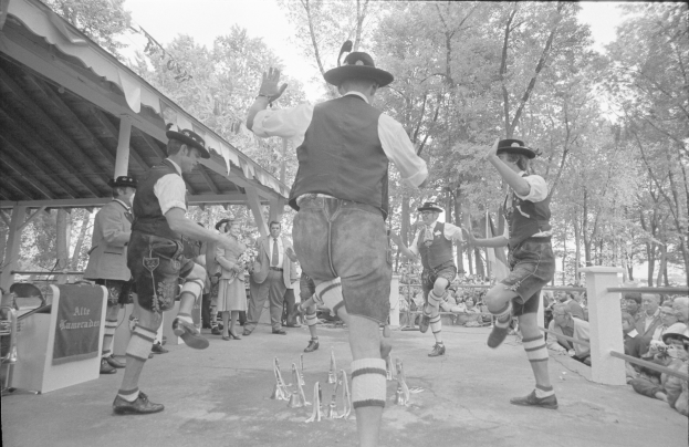 Eine Gruppe von Männern in traditioneller bayrischer Tracht tanzt auf einer Bühne vor einem gemischten Publikum aus sitzenden und stehenden Menschen, mit Bäumen und einer Hütte im Hintergrund, dargestellt in Schwarz-Weiß.