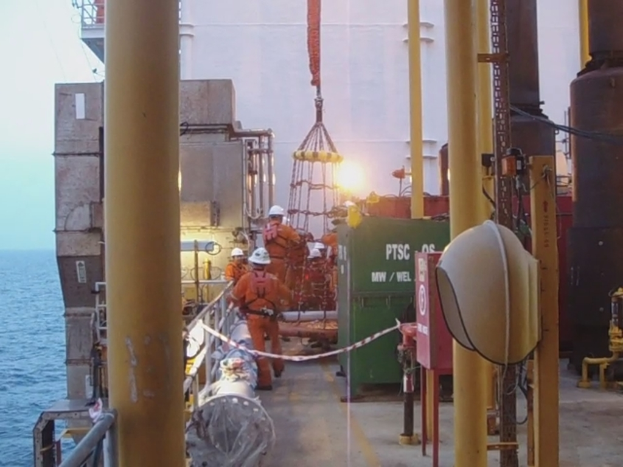 Ein Schiff mit Masten, Kisten, Geländern und Menschen mit Helmen, mit Wasser im Hintergrund.