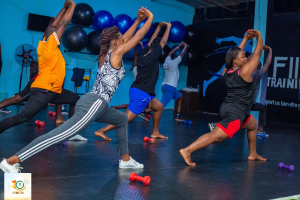 Gruppe von Menschen, die Yoga in einem Fitnessstudio machen, mit Hanteln auf dem Boden, einem Banner mit der Aufschrift "Fitness-Training in Ghana" im Hintergrund und diversen Gegenständen wie Luftballons und einem Ventilator.