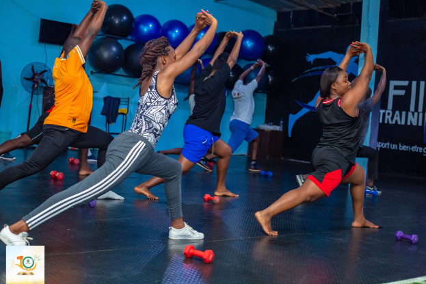 Gruppe von Menschen, die Yoga in einem Fitnessstudio machen, mit Hanteln auf dem Boden, einem Banner mit der Aufschrift "Fitness-Training in Ghana" im Hintergrund und diversen Gegenständen wie Luftballons und einem Ventilator.