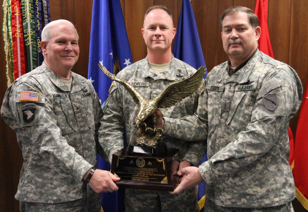 Drei Männer in Uniformen halten Auszeichnungen, mit Fahnen im Hintergrund und bunten Gegenständen mit Text in der oberen linken Ecke.
