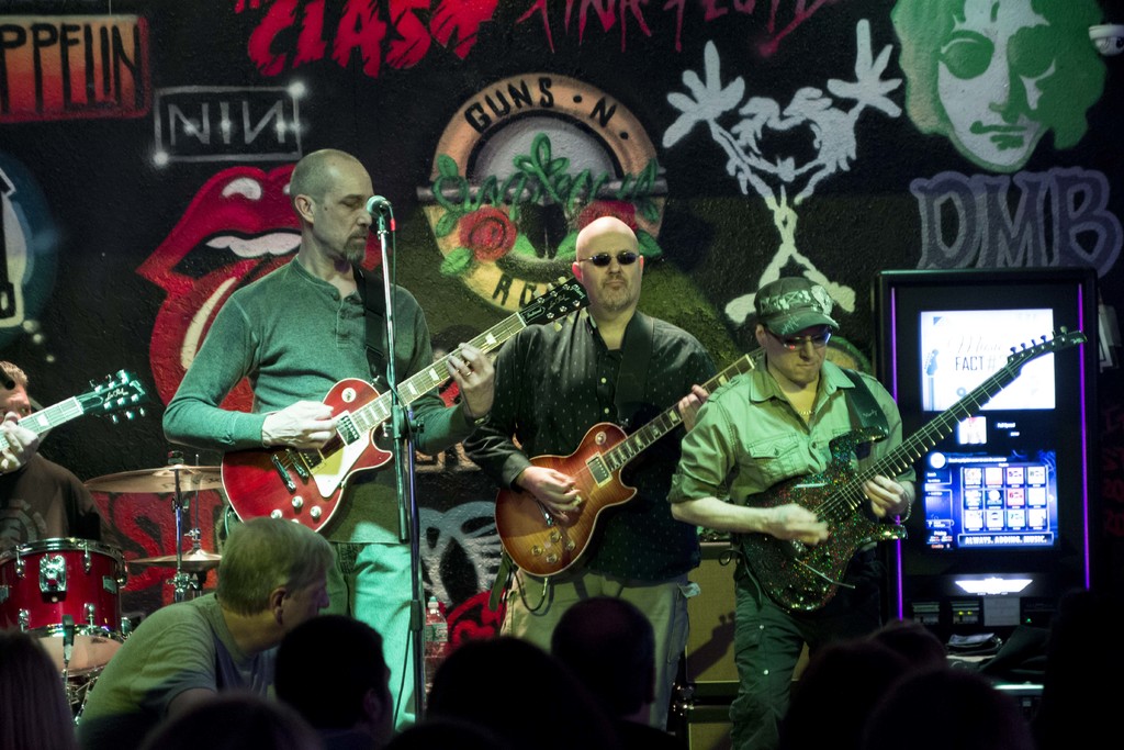 Drei Männer spielen Gitarren auf einer Bühne bei einem Konzert, mit einer Menge unten und einer gemalten Wand im Hintergrund.