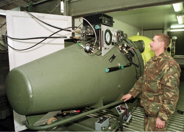 Ein Mann in militärischer Uniform steht neben einer großen grünen Bombe in einer unordentlichen industriellen Umgebung mit Maschinen, Drähten und einer beleuchteten Wand im Hintergrund, mit Text unten.