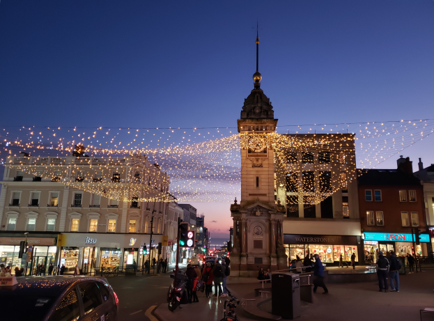 Eine belebte Stadtstraße bei Nacht mit Menschen, Fahrzeugen, Fahrrädern und Gebäuden, beleuchtet von Weihnachtslichtern, mit einem Uhrenturm im Hintergrund.