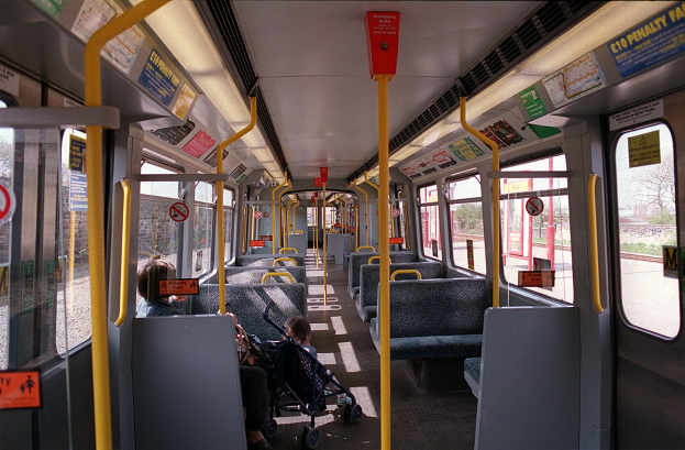 Innenseite eines öffentlichen Verkehrsmittels mit Sitzreihen, einem offenen Fenster und einem Baby im Kinderwagen, mit sichtbaren Hinweistafeln.