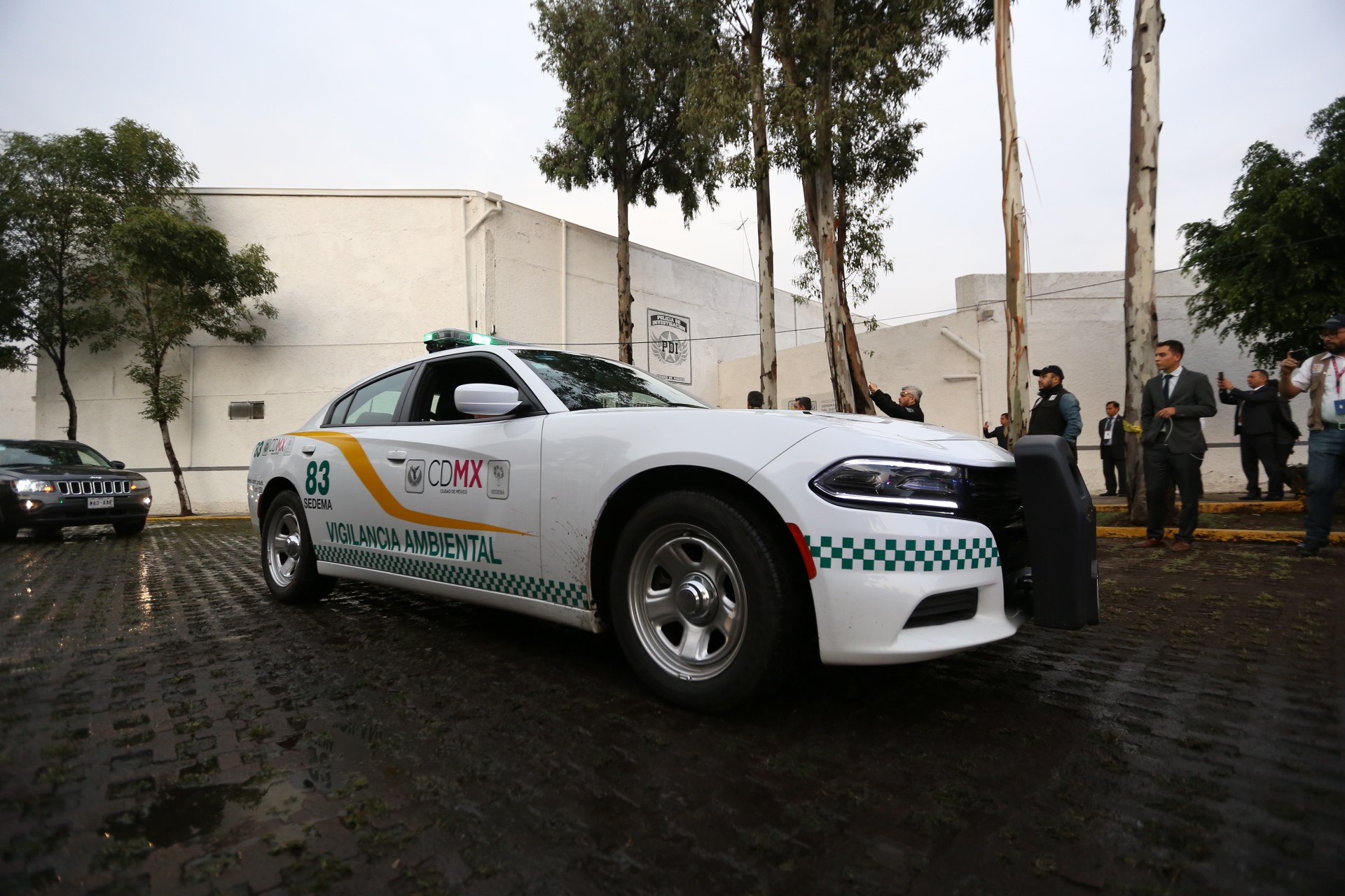 Weißer Polizeiwagen vor einem Gebäude mit einer Gruppe von Menschen in bunten Kleidern und Mützen, Bäumen und einem klaren blauen Himmel im Hintergrund.