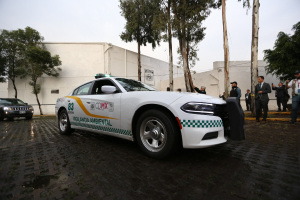 Weißer Polizeiwagen vor einem Gebäude mit einer Gruppe von Menschen in bunten Kleidern und Mützen, Bäumen und einem klaren blauen Himmel im Hintergrund.