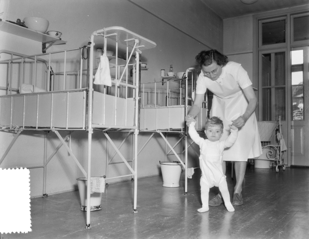 Ein Schwarz-Weiß-Foto einer Frau, die ein Baby in einem Krankenhauszimmer hält, mit einem Kinderbett, einem Eimer, einem Wagen, einer Tür, einem Fenster und Deckenleuchten.