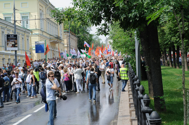Eine Gruppe von Menschen steht auf einer Straße außerhalb der Stadt, viele halten weiße und orangefarbene Fahnen, mit verschiedenen städtischen und natürlichen Elementen im Hintergrund.