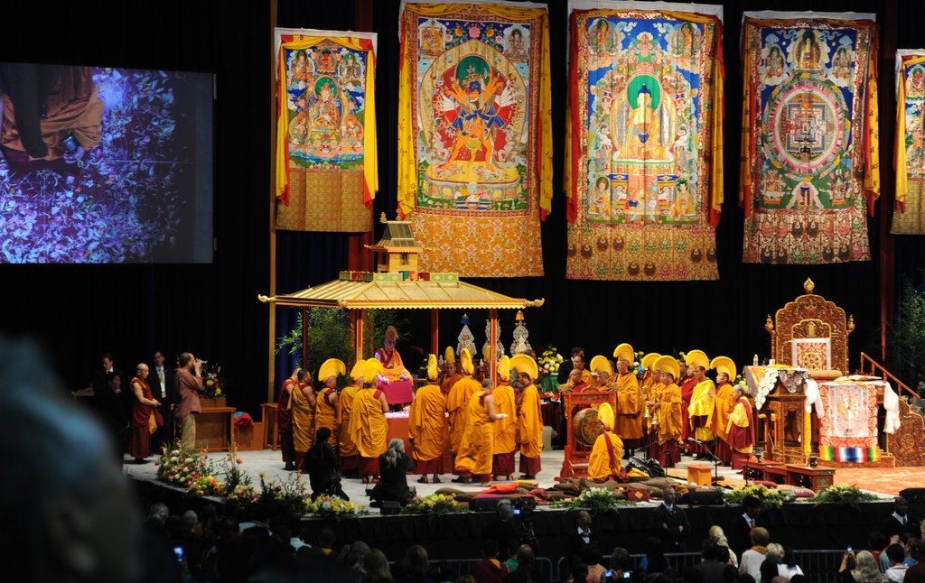 Eine Bühne mit Menschen, die performen, ein Bildschirm mit Inhalt, ein Schuppen im Hintergrund, Plakate einer Gottheit, Blumen und ein Publikum.