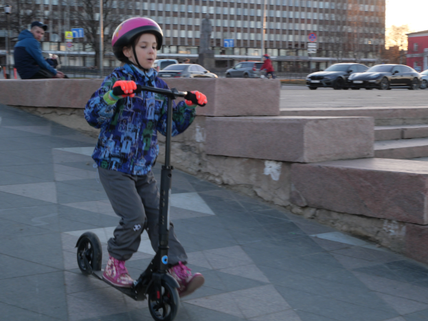 Ein junger Junge fährt mit einem Helm und Handschuhen auf einem Scooter auf einem Gehweg, mit verschiedenen städtischen Elementen und einem klaren blauen Himmel im Hintergrund.