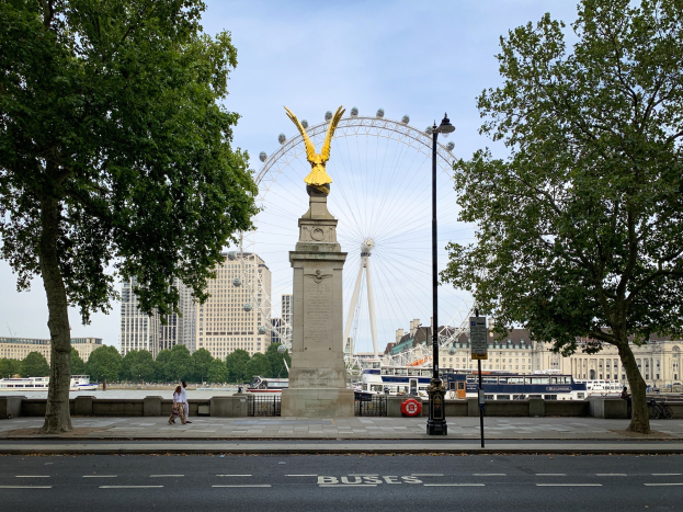 Das London Eye mit einer Statue auf einem Sockel im Vordergrund, Menschen auf einem Fußweg, Schiffe auf dem Wasser, Bäume, Gebäude und der Himmel.