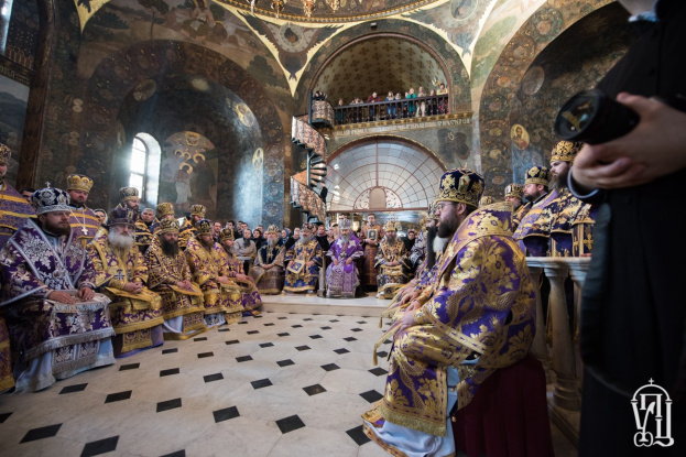 Eine Gruppe von Menschen in einer Kirche, einige sitzen und andere stehen auf einer Treppe, mit einer Person, die rechts eine Kamera hält; ein Kronleuchter hängt von der Decke, und Gemälde schmücken die Wände.