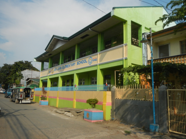 Ein Schulgebäude mit einer grünen und gelben Fassade, das neben einer Straße mit Fahrzeugen, Topfpflanzen, einem Zaun, einem Tor, einem Straßenschild, Bäumen, Stromleitungen und einem bewölkten Himmel steht.