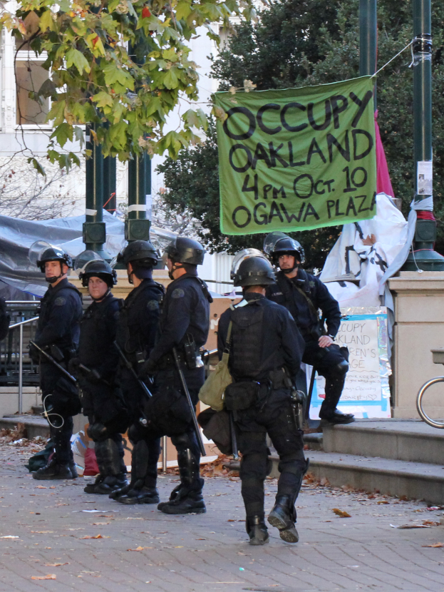 Eine Gruppe von Polizisten in Helmen, die Waffen halten, geht eine von Bäumen gesäumte Straße entlang mit verschiedenen Gegenständen und einem Gebäude im Hintergrund.