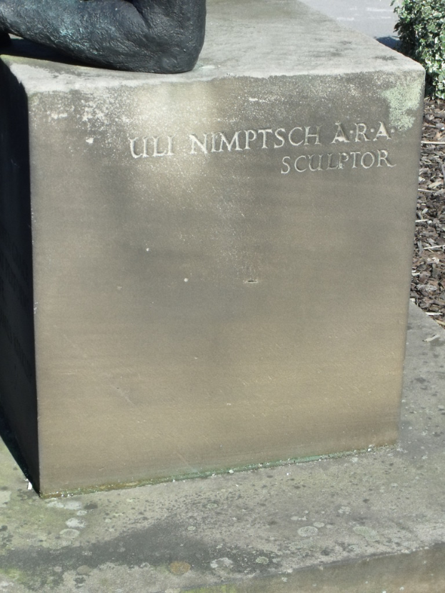 Ein Stein mit Text in Vordergrund, eventuell eine Statue oben, trockene Zweige, Erde und Pflanzen rechts, mit einer Straße im Hintergrund.