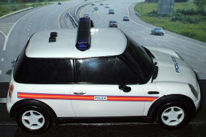 Weißer Polizeiwagen mit "Polizei" darauf auf einer Straße außerhalb der Stadt fahrend, umgeben von fließendem Verkehr, Gras und Bäumen.