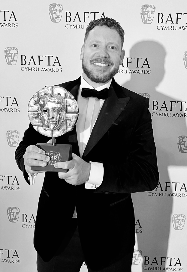 Ein Mann in einem Smoking, der eine BAFTA-Trophäe hält und lächelt, mit einem Hintergrundplakat, auf dem 'BAFTA Awards 2018' steht