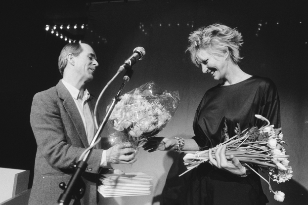 Ein Mann im Anzug und eine Frau mit einem Blumenstrauß stehen zusammen auf einer Bühne, lächeln und haben ein Mikrofon vor sich und verstreute Papiere, beleuchtet von Scheinwerfern vor einem dunklen Hintergrund.