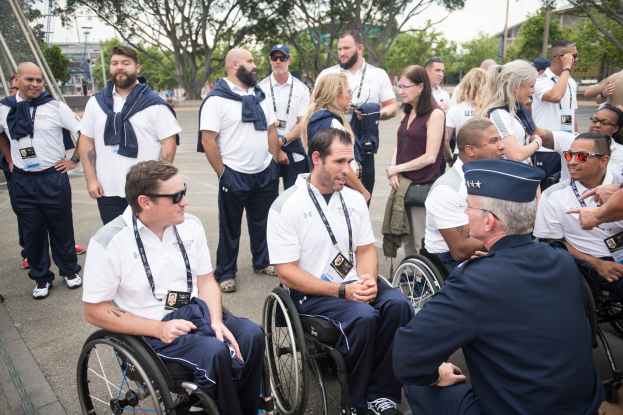 Eine Gruppe von Menschen im Rollstuhl spricht vor einer Kulisse mit Bäumen, Pfählen, Gebäuden und einem klaren blauen Himmel, wobei einige Mützen und Brillen tragen, was auf ihre Teilnahme an den Paralympischen Spielen hinweist.