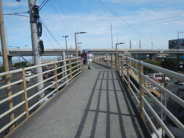 Eine Fußgängerbrücke mit Menschen, die überqueren, umgeben von Geländern, Laternen, Strommästen mit Kabeln, Fahrzeugen auf der Straße, Bäumen, Gebäuden und einem bewölkten Himmel im Hintergrund.