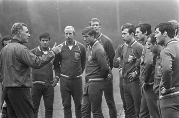 A black and white photo of a group of men standing on a soccer field, with the man in the center wearing a walking shoe.
