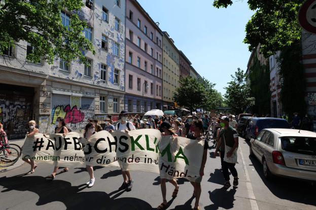 Eine Gruppe von Menschen marschiert auf einer Straße mit einem Banner, rechts parkende Autos, links ein Fahrradfahrer, Gebäude und Bäume im Hintergrund und eine Berliner Demonstration im Hintergrund.