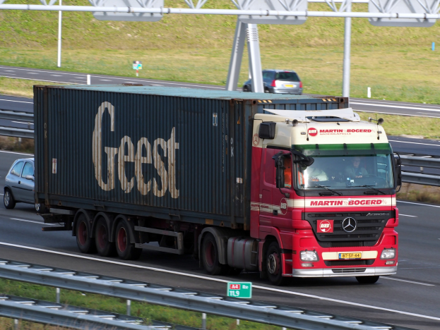 Roter Lastwagen mit einem Container auf einer Autobahn unterwegs, mit Passagieren, Strommasten, Holzplanken und Gras im Hintergrund.