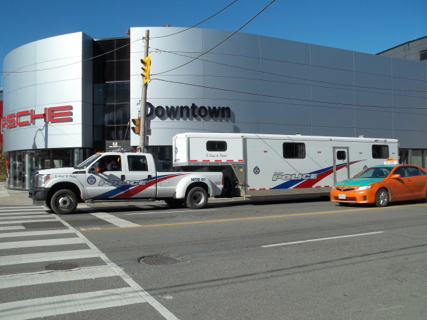Ein Polizei-Lkw vor einem Gebäude mit Text darauf geparkt, mit einer Ampel mit Stange und Kabeln im Vordergrund und Bäumen und einem klaren blauen Himmel im Hintergrund.