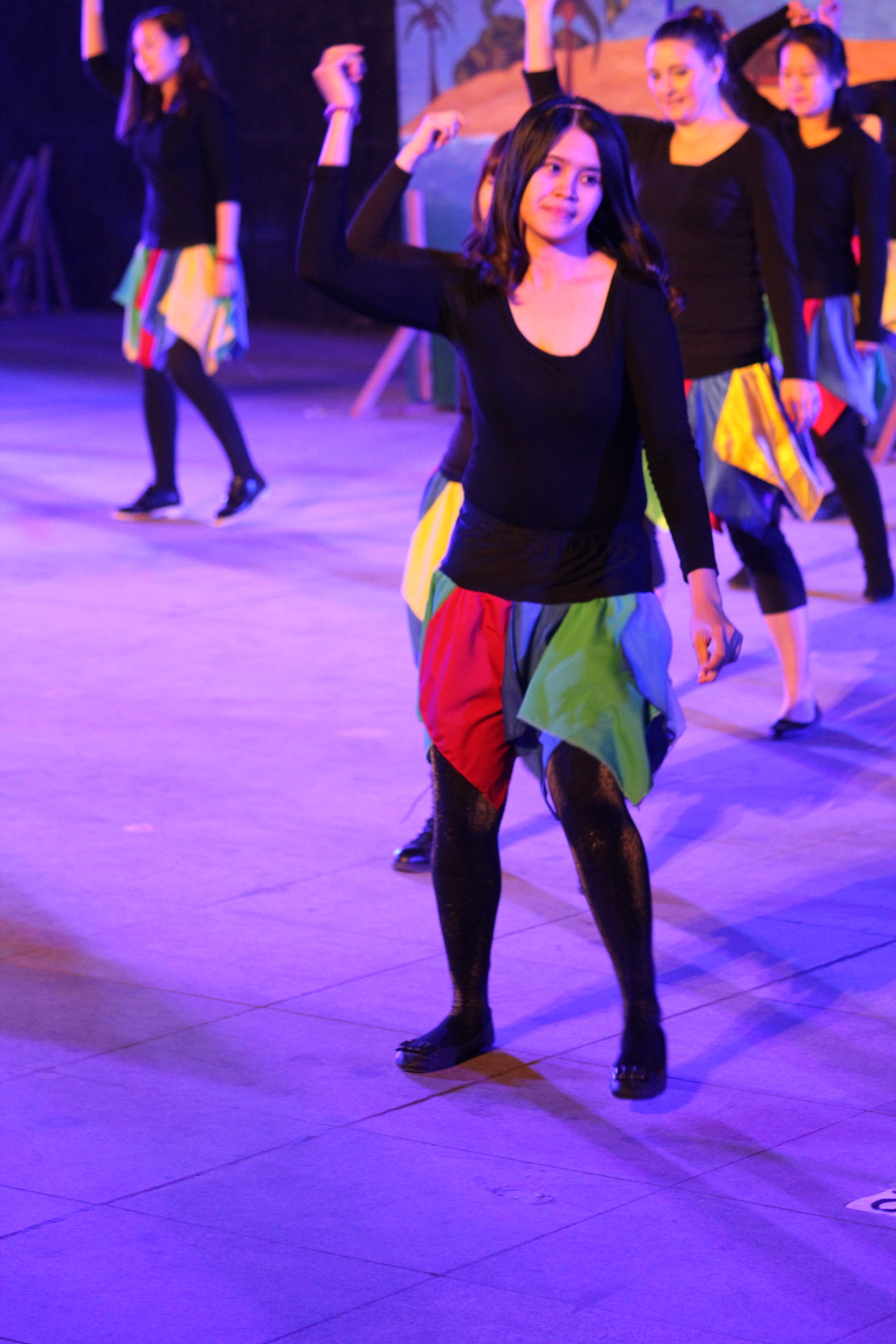 Eine Gruppe junger Frauen, die auf einer Bühne tanzen, mit einem Gemälde im Hintergrund, alle tragen Schuhe.