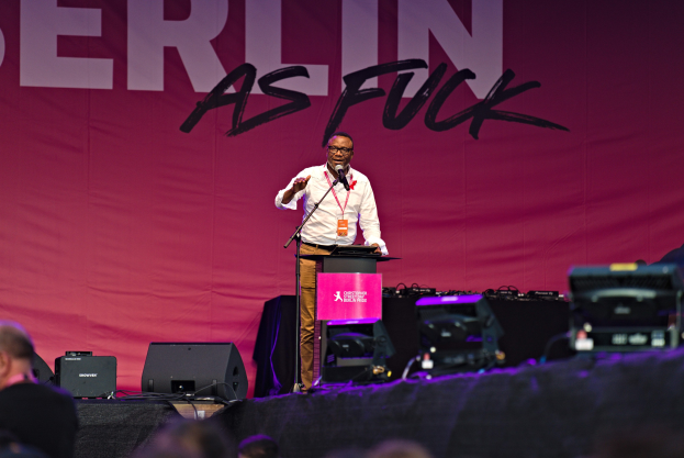 Ein Mann in einem weißen Hemd und braunen Hosen steht auf einer Bühne und hält ein Mikrofon in der Hand, während er eine Menge anspricht. Im Hintergrund hängt ein pinkfarbenes Banner mit der Aufschrift 'Berlin as Fuck'.