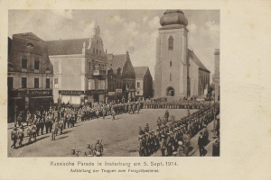 Schwarzes und weißes Foto einer Parade in Insterburg 1914 mit Menschen und Gebäuden im Hintergrund, Wolken am Himmel und Text unten.