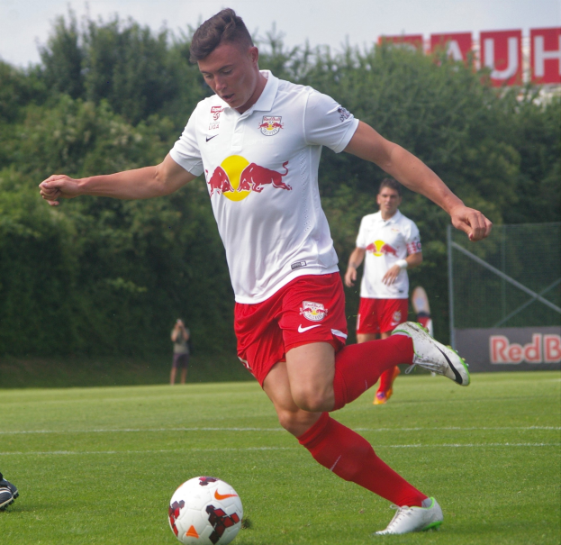 Ein Mann in einem Fußballtrikot kickt einen Ball auf einem grünen Feld mit Bäumen und einem klaren blauen Himmel im Hintergrund, mit einem Schild, auf dem "RB Leipzig" zu lesen ist.